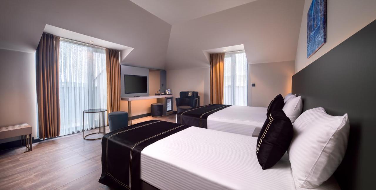 Menalo Hotel Premium Istanbul Airport Zewnętrze zdjęcie The photo shows a modern hotel room featuring two beds with white linens and black throw pillows. There is a television mounted on the wall opposite the beds. A small seating area includes a comfortable chair and a table, while a mini-fridge and a co
