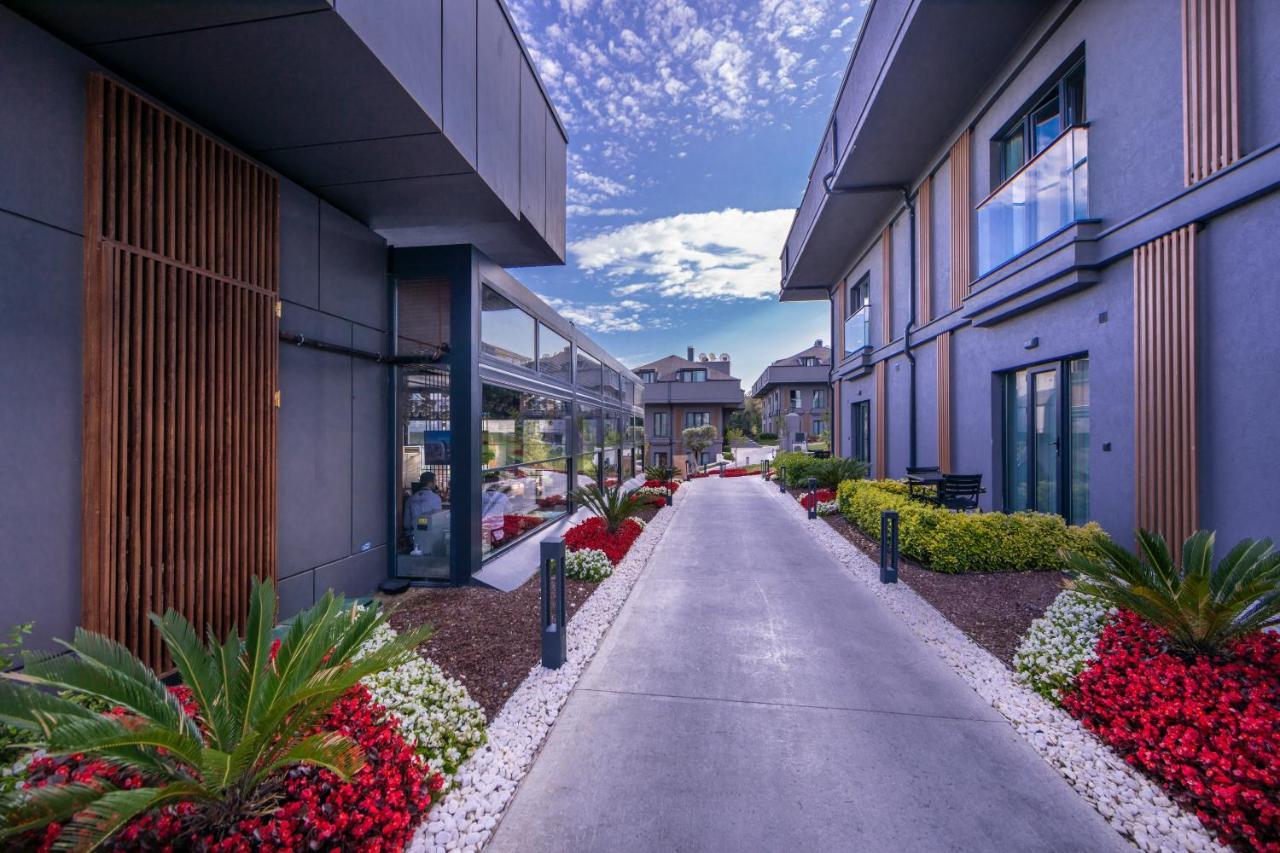 Menalo Hotel Premium Istanbul Airport Zewnętrze zdjęcie The photo depicts a well-designed exterior pathway leading between modern buildings. The pathway is bordered by lush greenery and vibrant flower beds featuring a mix of red, white, and green plants. The sky above is partly cloudy, adding a serene bac