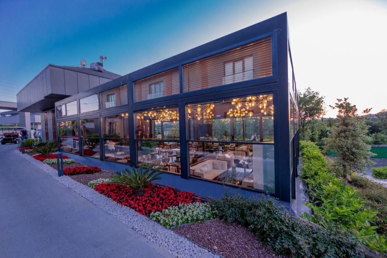Menalo Hotel Premium Istanbul Airport Zewnętrze zdjęcie The photo shows a modern restaurant or dining space characterized by large glass windows that create an open and inviting atmosphere. The exterior features a sleek, dark frame with a contemporary design. There are vibrant flower beds with red and gre