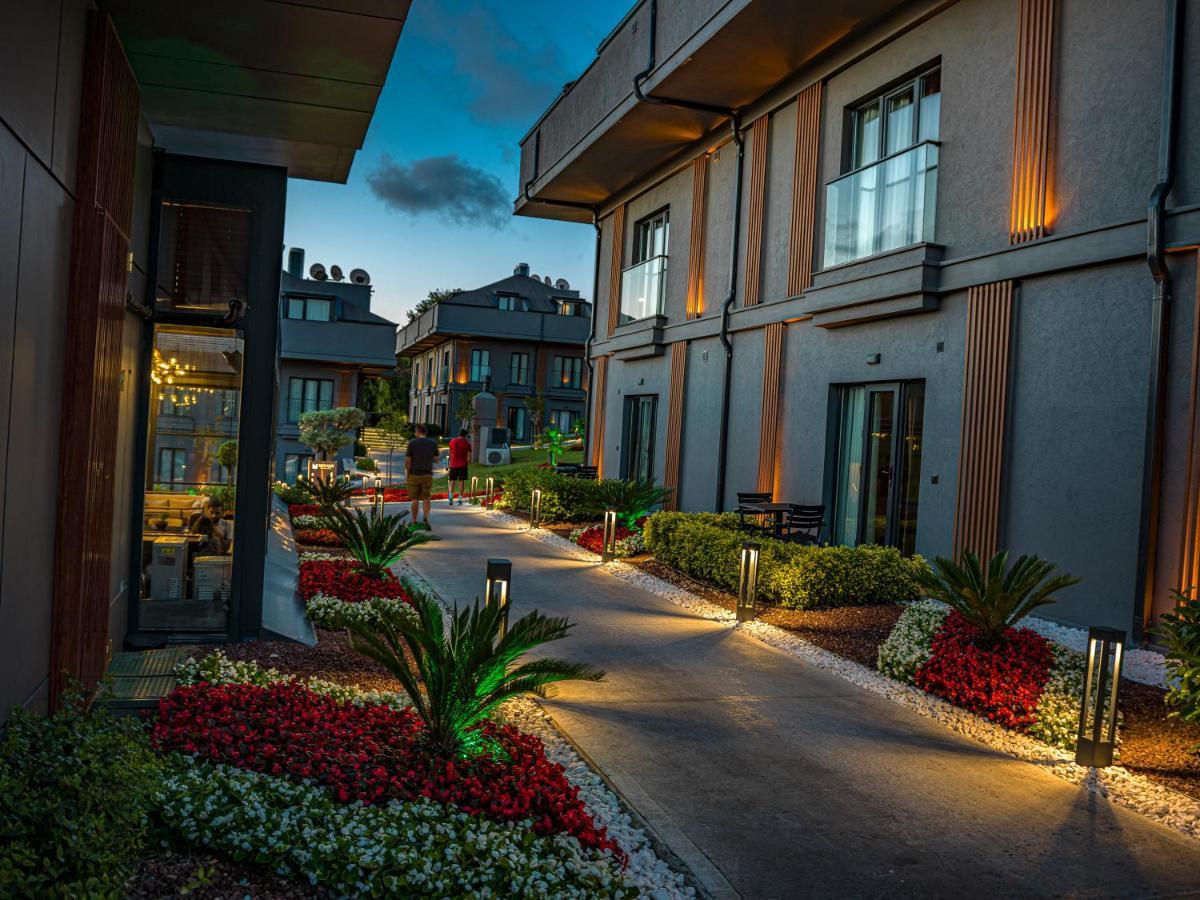 Menalo Hotel Premium Istanbul Airport Zewnętrze zdjęcie The photo shows a pathway between modern buildings, illuminated by soft lighting. The path is lined with colorful flower beds featuring red and white blooms and small palm plants, adding to the aesthetic appeal. The buildings are designed with a mix 