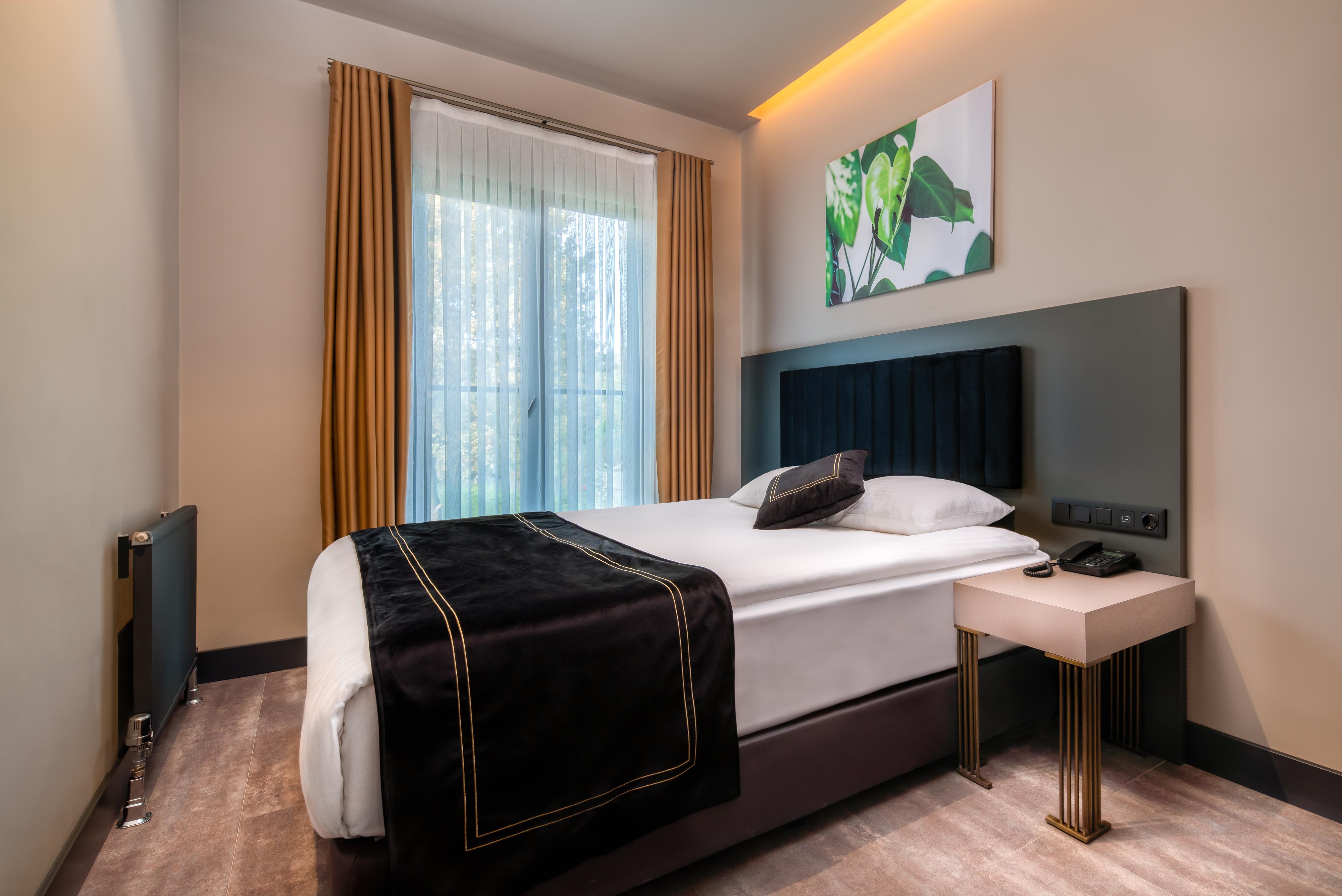 Menalo Hotel Premium Istanbul Airport Zewnętrze zdjęcie The photo shows a modern hotel room featuring a neatly made bed with a black and white bedspread. The headboard is upholstered in a dark fabric. There's a small bedside table beside the bed with a lamp or phone. The room has large windows covered wit