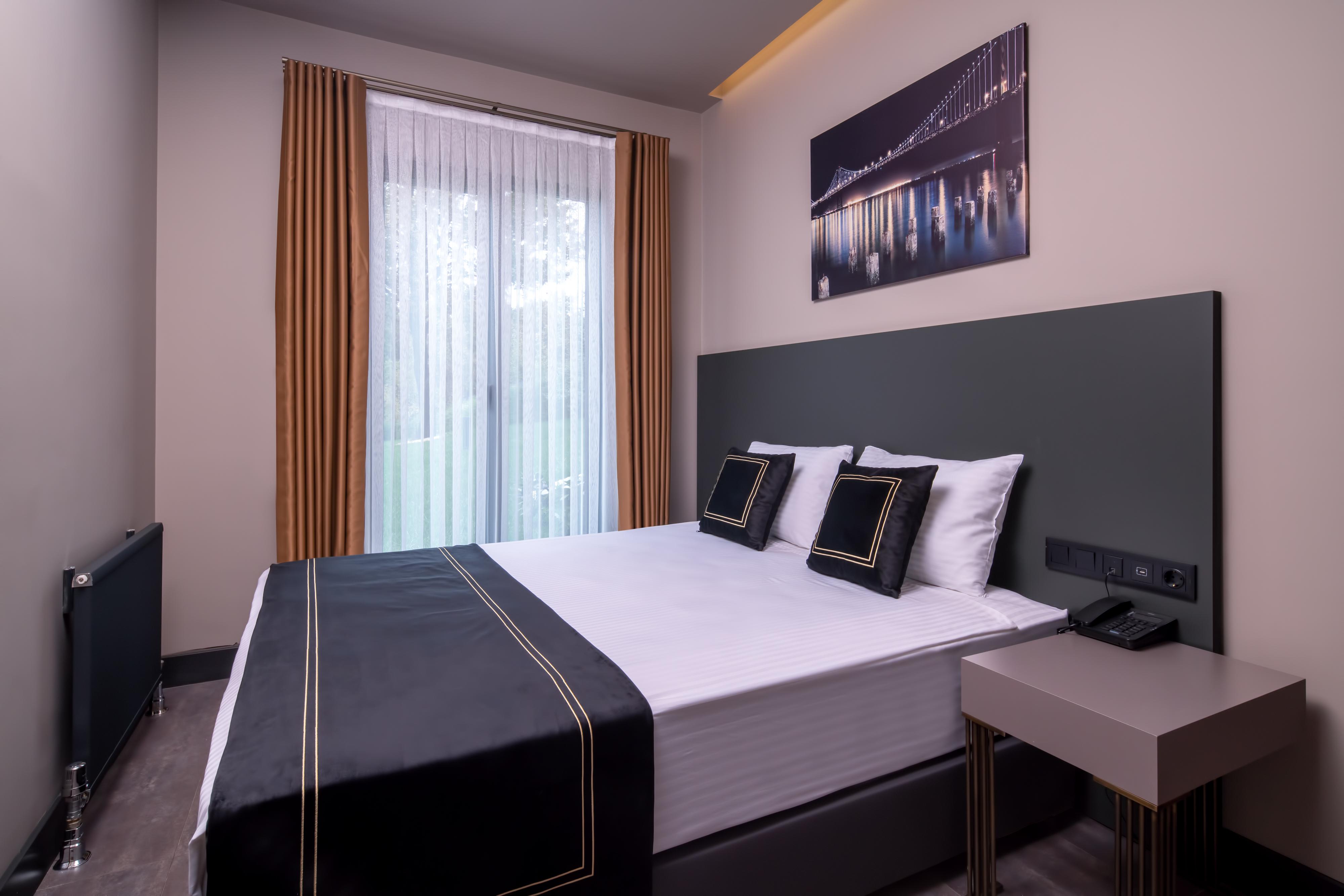 Menalo Hotel Premium Istanbul Airport Zewnętrze zdjęcie The photo shows a modern hotel room featuring a double bed with a sleek black and white bedding set. Two decorative pillows are placed on the bed. There are large windows with sheer curtains that allow natural light to enter the room, and the walls a