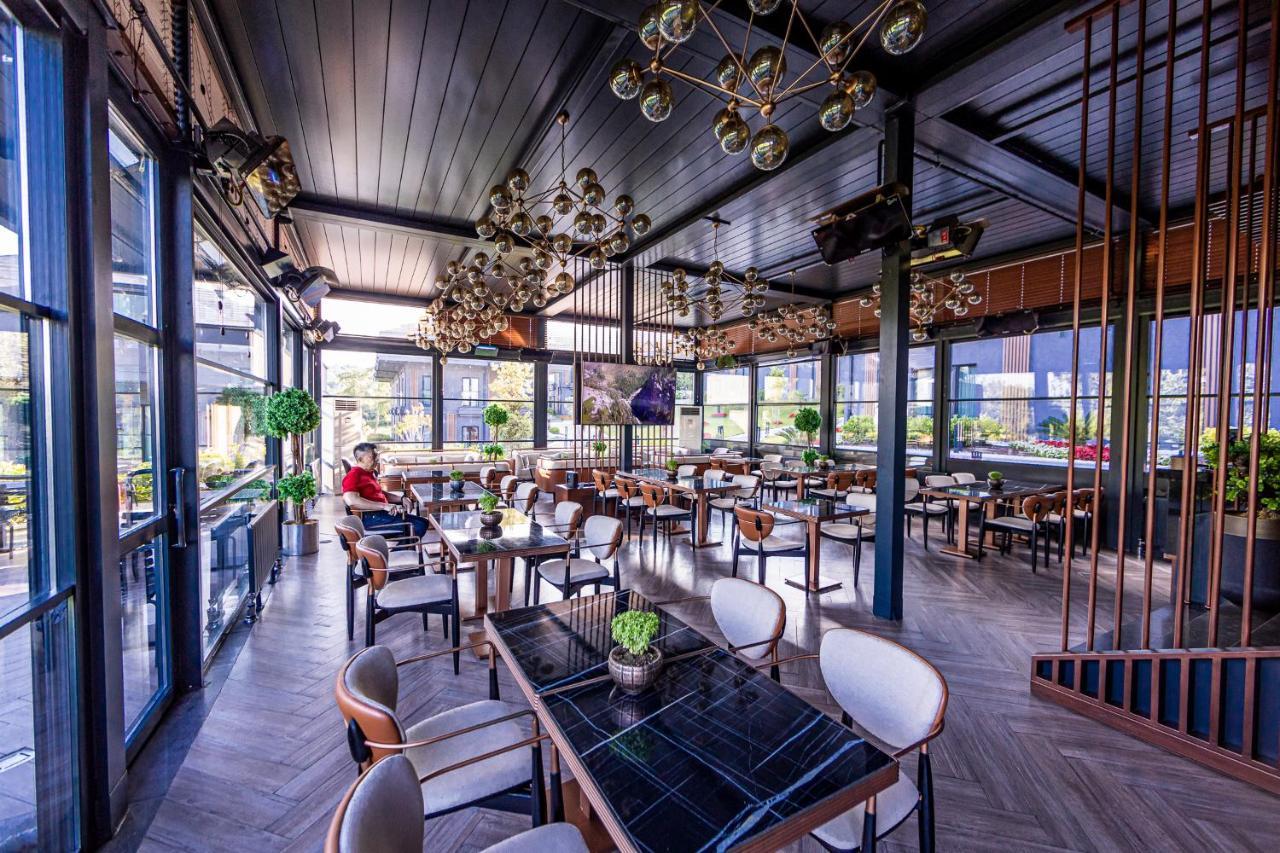 Menalo Hotel Premium Istanbul Airport Zewnętrze zdjęcie The photo shows a stylish restaurant interior that features modern decor. There are several tables arranged around the space, each with elegant seating. The tables are set with small green plants as centerpieces. The ceiling is adorned with decorativ