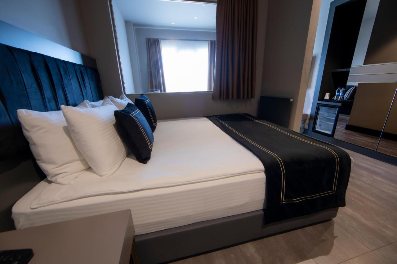 Menalo Hotel Premium Istanbul Airport Zewnętrze zdjęcie The photo shows a modern hotel room featuring a queen-sized bed with a dark headboard. The bed is dressed with white and dark-colored pillows, and there is a black blanket draped over the foot of the bed. A window with curtains allows natural light t
