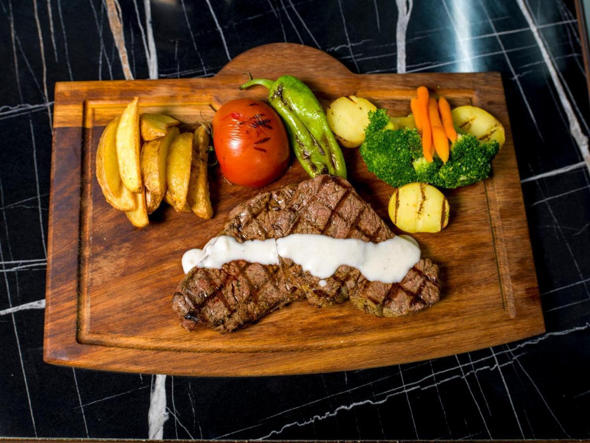 Menalo Hotel Premium Istanbul Airport Zewnętrze zdjęcie The photo shows a meal presented on a wooden cutting board. The main feature is a grilled steak, which has grill marks and is topped with a creamy sauce. Accompanying the steak are several side items: 

- A portion of golden-brown potato wedges
- A w