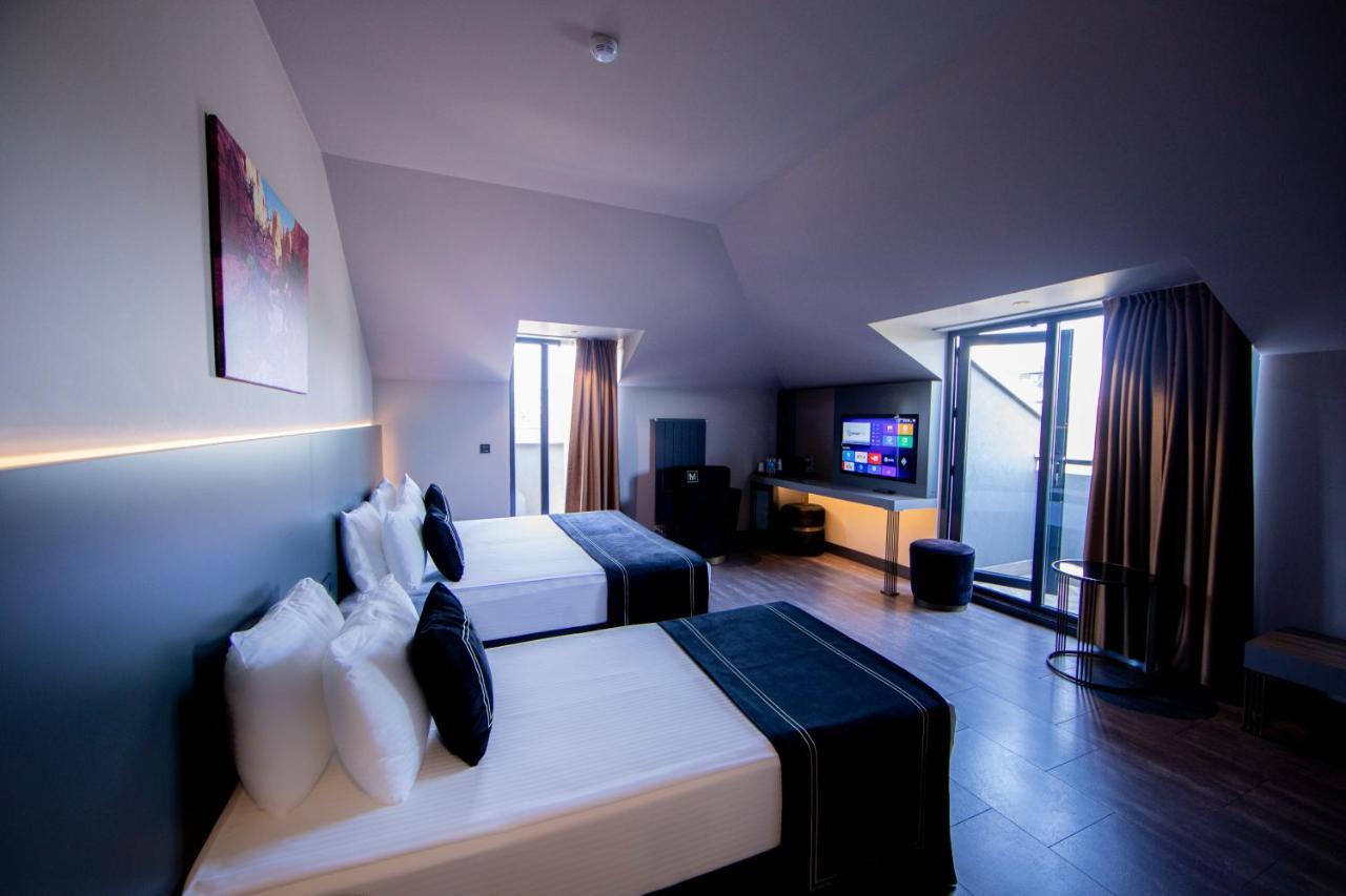 Menalo Hotel Premium Istanbul Airport Zewnętrze zdjęcie The photo shows a modern hotel room with a sleek design. There are two beds with white linens and dark blue accents. A large flat-screen TV is mounted on the wall, and there is a small seating area with a chair and a table. The room features large wi